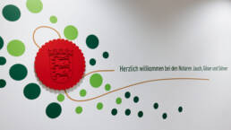 Wandgestaltung mit rotem Wappen und dem Schriftzug: „Herzlich willkommen bei den Notaren Jauch, Göser und Söhner“.