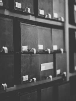 Regalwand mit alten, dunklen Aktenboxen, Metallgriffen und handschriftlichen Etiketten.