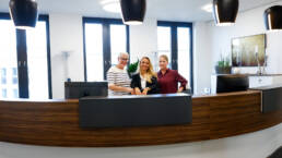 Drei freundliche Mitarbeiterinnen stehen lächelnd hinter einer geschwungenen hölzernen Empfangstheke.